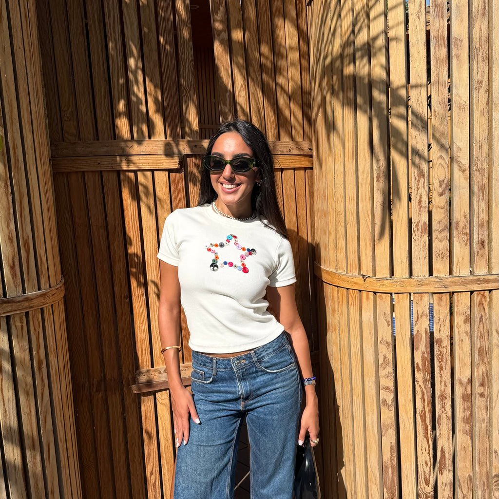 A woman wearing a white crewneck t-shirt with a colorful floral graphic on the chest and blue wide-leg jeans, standing in front of a wooden slatted wall.