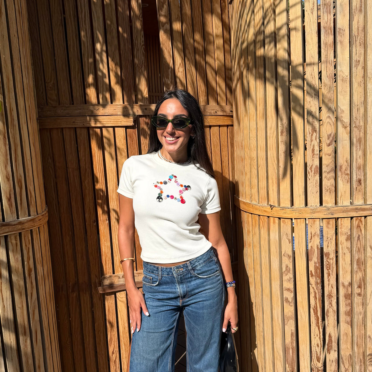 A woman wearing a white crewneck t-shirt with a colorful floral graphic on the chest and blue wide-leg jeans, standing in front of a wooden slatted wall.